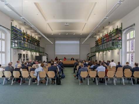 Blick in einen Raum mit vielen Stühlen. Auf denen sitzen Menschen. An einem Tisch unter einer Leinwand sitzen drei Menschen. An den Wänden sind Bücherregale. 