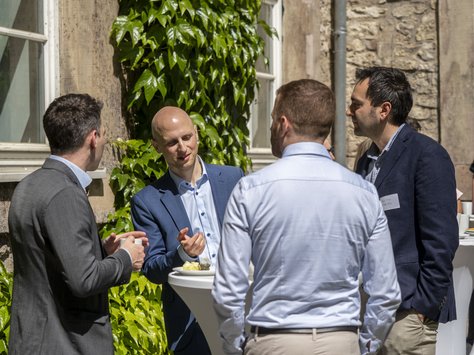 Vier Männer in Businesskleidung stehen an einem Stehtisch. Im Hintergrund ist eine Hausfassade mit Fenstern und Pflanzenbewuchs zu sehen. 