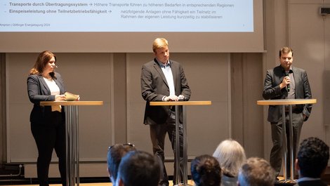In einem Tagungsraum stehen eine Frau und zwei Männern an Stehtischen. Der Mann rechts hält ein Mikrofon in der Hand.