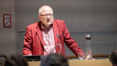 Ein Mann mit rotem Sakko und Brille steht an einem Rednerpult.