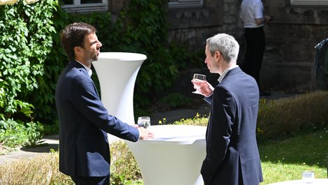 Zwei MMänner in Businesskelidung stehen an einem Stehtisch, der mit einer weißen Husse überzogen ist. der rechte Mann hat ein Glas in der Hand. Sie stehen in einem begrünten Innenhof.