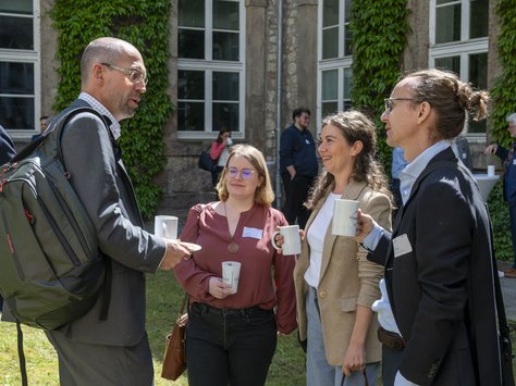 Drei Frauen und ein mann mit Rucksack stehen vor einer Fassade mit Fenstern. 