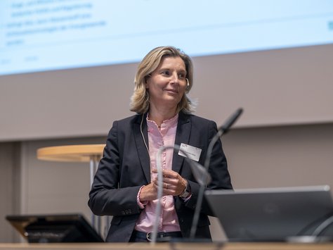 Eine blonde Frau steht an einem Mikrofon vor einer Leinwand.