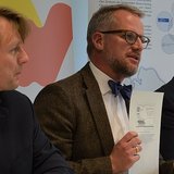 drei Personen sitzen nebeneinander, eine Person hält ein Blatt Papier