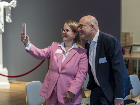 Eine Frau mit einer rosa Jacke und ein Mann in weißem Hemd und dunklem Sakko stehen in einem Innenraum und machen ein Handy-Selfie. 