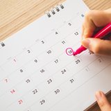 Eine Hand kreist auf einem Monatskalender ein Datum mit einem roten Stift ein. 