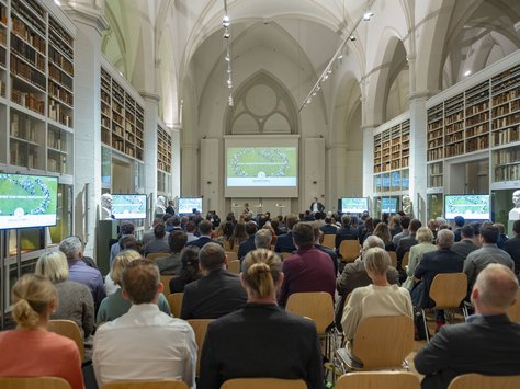 Blick in einen Tagungsraum. Der Raum hat ein Kreuzrippengewölbe und gotisch anmutende Bögen.  An den Wänden befinden sich Bücherregale. Der Raum ist mit Menschen auf Stühlen gut gefüllt. An den Seiten stehen Bildschirme, an der Front eine Leinwand. 