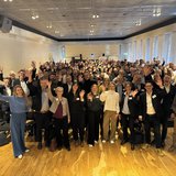 Zahlreiche Menschen stehen in einem Raum und winken.