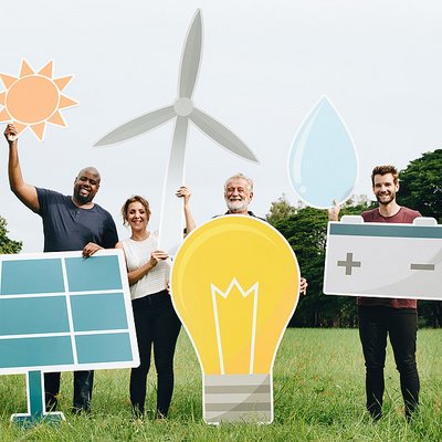 Vier Personen stehen im Gras und halten einen Papierausschnitt eines Objekts. Eine hält eine Glühbirne, eine hält eine Batterie und ein Wassersymbol, eine hält ein Solarpanel, eine hält ein Windrad