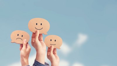 Mehrere Hände halten Sprechblasen aus Papier mit freudigen, neutralen und traurigen Gesichtern darauf hoch