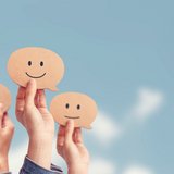 Mehrere Hände halten Sprechblasen aus Papier mit freudigen, neutralen und traurigen Gesichtern darauf hoch
