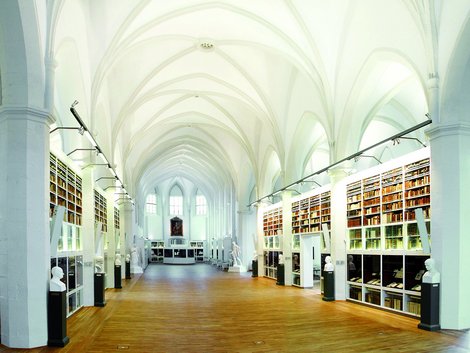 Ein Hallenartiger Inneraum mit Kreuzgewölbe und Bücherragalen an den Wänden.